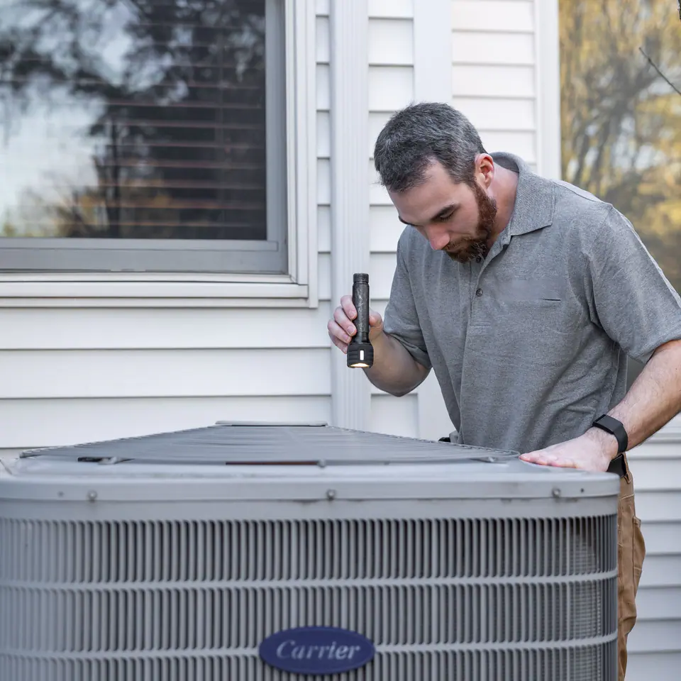 A technician inspecting an outdoor air conditioning condenser beside a white house