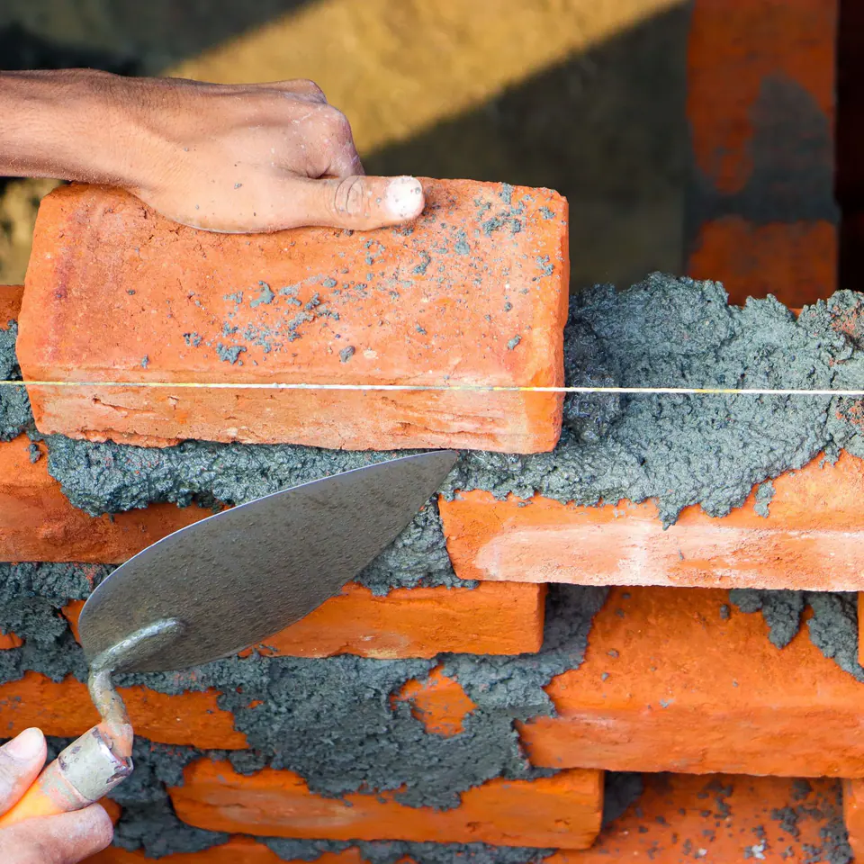 Hands laying an orange brick on fresh mortar with a trowel, a string line across the wall