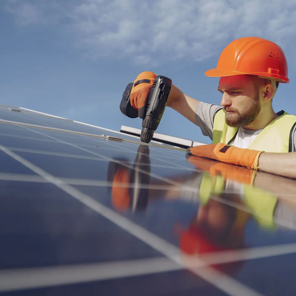An installer in an orange hard hat fixing solar panels on a roof with a power drill