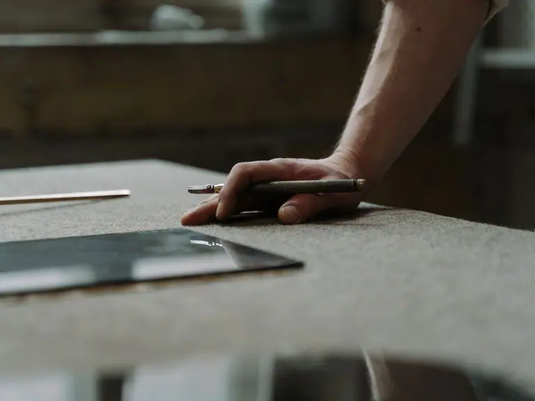 A hand resting beside a glass cutter and a cut pane of glass on a felt-topped workbench