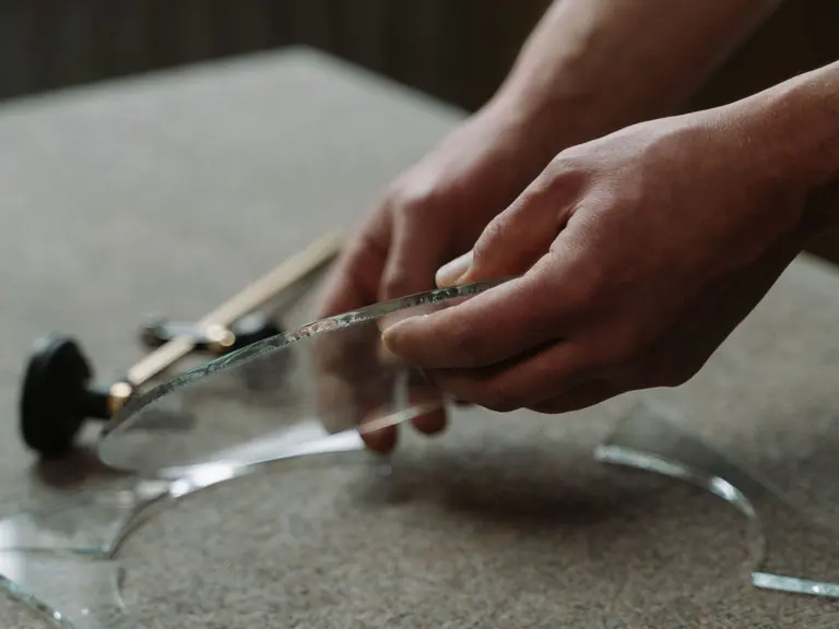 Two hands snapping a scored glass circle free of its waste piece, a glass cutter resting beside it