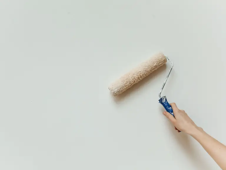 A hand holding a paint roller loaded with pale paint against a plain white wall