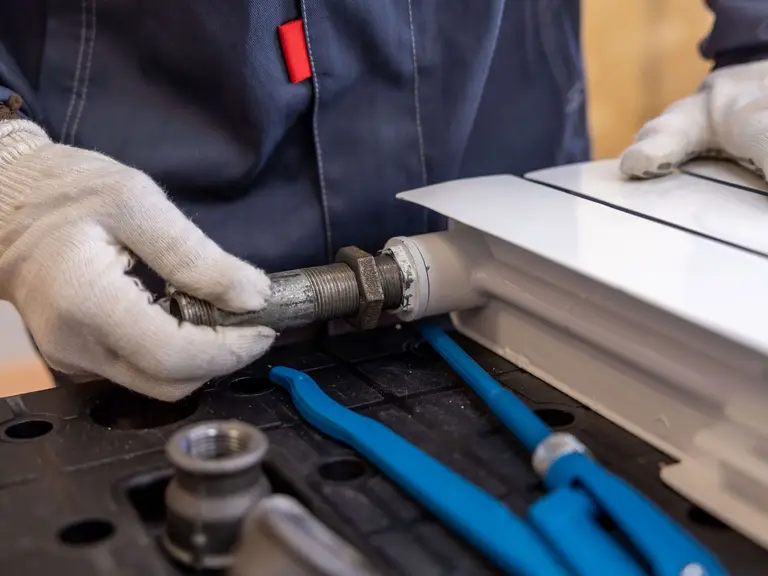 A plumber's gloved hands threading a steel pipe fitting into a radiator connection
