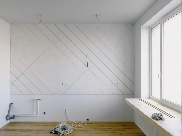An empty room mid-renovation with rough-in plumbing and electrical wiring exposed against a wall marked out for tile