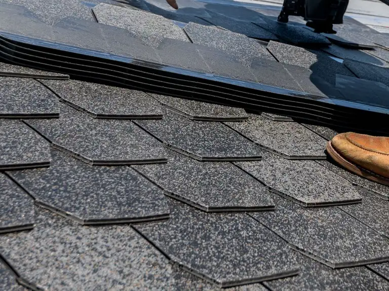 A roofer bracing a row of asphalt shingles with a boot beside a pneumatic nail gun on a pitched roof