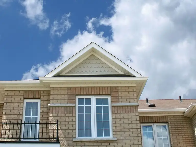 The brick upper facade of a suburban house with a tall paned picture window, a French-door balcony and a smaller window, under a partly cloudy sky